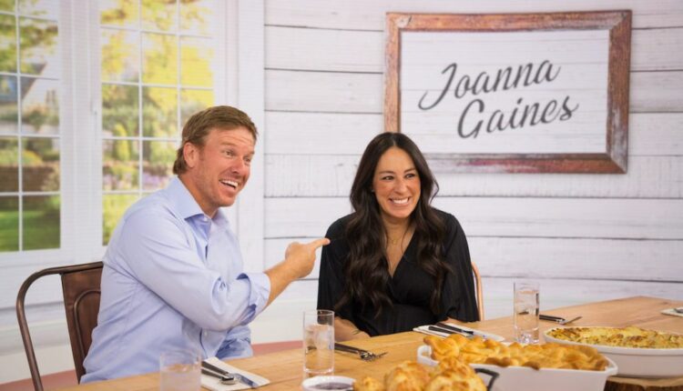 Chip smiles and points to Joanna Gaines while the two sit at a table