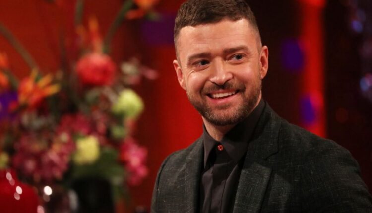 Justin Timberlake, known to an average car, dressed in a black suit with a black shirt in front of a red background with a floral arrangement off to the side.